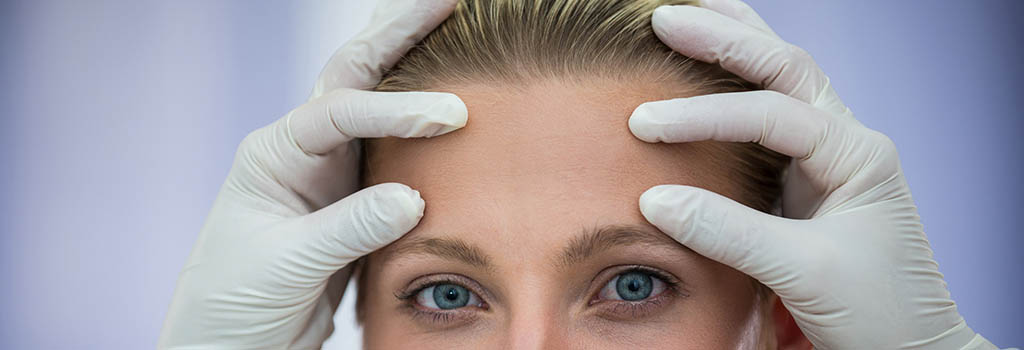 Na imagem aparece o rosto de uma mulher, com close na testa e na região dos olhos, e as mãos de um médico usando luvas cirúrgicas com os dedos polegares e indicadores posicionados na testa, simulando uma consulta. 