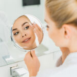 A imagem mostra uma mulher caucasiana, com cabelos loiros presos em um coque, de costas, se olhando em um espelho redondo. À frente da modelo, está outro espelho, que toma toda a parede. Abaixo dele, há uma pia com torneira prateada.