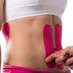 Um close no abdômen de uma mulher branca, uma mão coloca bandagens adesivas de cor rosa em sua barriga, no sentido vertical. A imagem tem fundo branco.